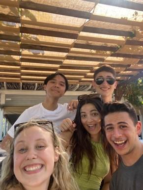 Jack en vier andere jongeren nemen een selfie. Ze dragen zomerkleren en staan ergens buiten onder een afdak waar de zon door komt.