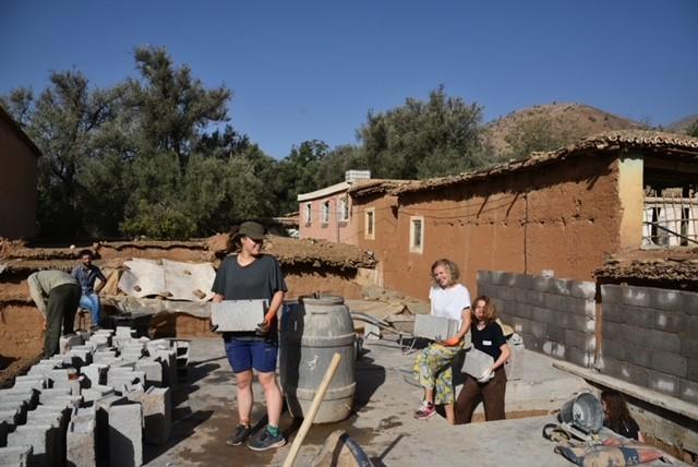 Esther met twee andere vrijwilligers tijdens het metsen van een muur in Marokko. Met op de achtergrond een gebouwtje en bomen, tegen een blauwe hemel.