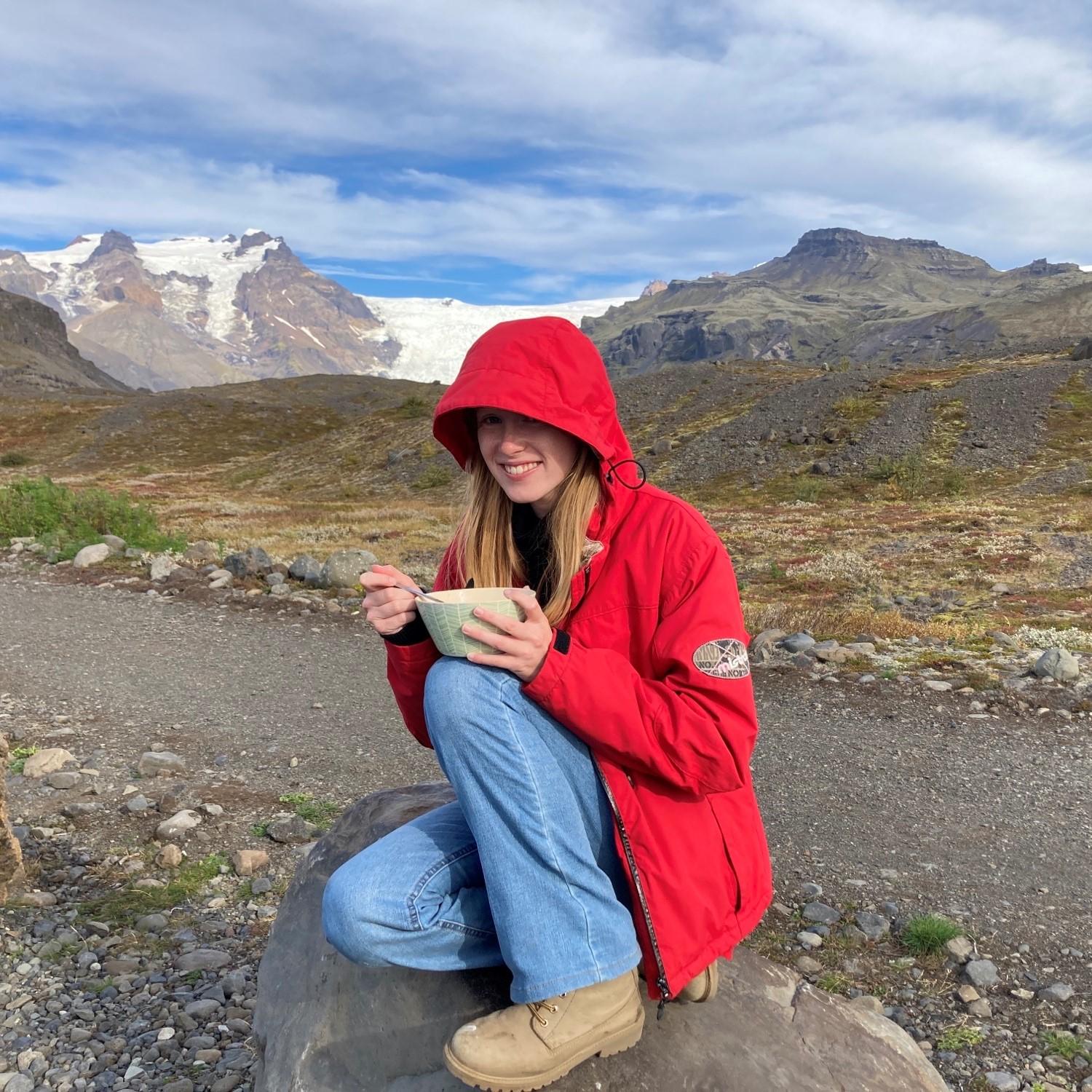 Fien in IJsland lachend naar de foto met een rode regenjas. Ze zit op een rotsblok en eet iets uit een kommetje